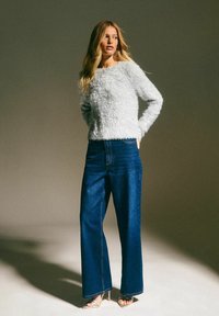 Puszysty jasnoszary sweter z okrągłym dekoltem zestawiony z ciemnoniebieskimi spodniami o wysokim stanie i szerokimi nogawkami oraz srebrnymi sandałami na obcasie z odkrytymi palcami.