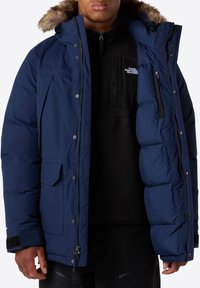 Giacca puffer blu navy con cappuccio foderato in pelliccia, dotata di zip frontale, bottoni a pressione e numerose tasche. Strato interno testurizzato visibile.