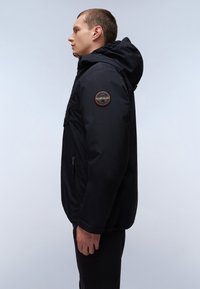 Schwarze Kapuzenjacke mit einem Reißverschluss, ausgestattet mit Seitentaschen und einem runden Logopatch am linken Arm. Glatte Textur und figurbetontes Design.