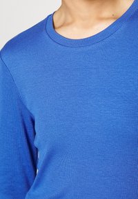 Modré žebrované tričko s dlouhými rukávy, kulatým výstřihem, hladkou texturou a přiléhavým střihem, zdůrazňující živou barvu a rovnoměrný vzor.