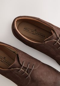 Paire de chaussures en cuir marron à lacets avec semelles intérieures beiges imprimées « Hell for Leather Next » sur fond blanc.