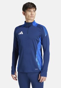 T-shirt de sport à manches longues bleu marine avec des accents bleus, col zippé à mi-hauteur et logo Adidas. Texture lisse avec un motif graphique sur le côté.