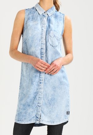 Sleeveless light blue acid-wash denim shirt dress with collar, front buttons, chest pocket, and side slit worn over black pants.