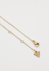 Gouden ketting met een driehoekige hanger waarop "GUESS" is gegraveerd. Het is ontworpen met een kreeftenhaak voor een veilige sluiting.