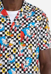 Kurzarmhemd mit einem bunten, animierten Sonic the Hedgehog-Print auf einem schwarz-weißen karierten Hintergrund. Das Material scheint aus Baumwolle zu sein.