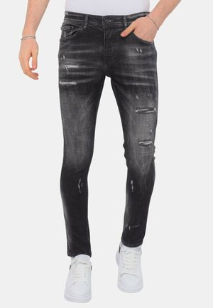 Man draagt zwarte slim-fit versleten jeans en witte sneakers met één hand in de zak tegen een effen achtergrond.