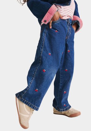 Enfant portant un jean bleu large avec une broderie de cerise rouge, des baskets beige, un t-shirt à rayures et une veste en denim aux poignets retroussés.