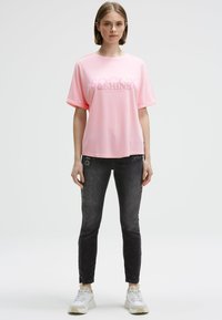 Young woman stands facing forward wearing a loose pink T-shirt with graphics, black slim jeans, and white sneakers on a plain background.