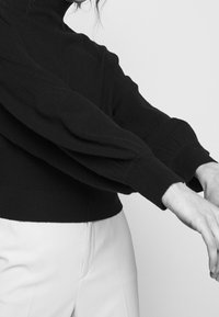 Svart stickad tröja med hög krage och voluminösa ärmar, ribbade muddarna och en figursydd midja, i kombination med ljusa byxor.