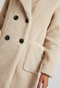 Abrigo de vellón beige con diseño de doble botonadura, botones negros, grandes bolsillos frontales y tejido texturizado para mayor abrigo.