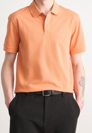 Mann trägt ein kurzärmliges oranges Polo-Shirt mit „BOSS“-Logo und schwarze Hose, Hände in den Taschen, zeigt ein Tattoo am linken Unterarm.