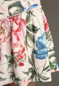 Kukkakuosiset mekko, jossa on vaaleanpunaisia, punaisia ja sinisiä kukkia tekstuuripinnalla. Puseroitu helma vihreillä lehdillä, pehmeä pinta ja vaalea tausta.