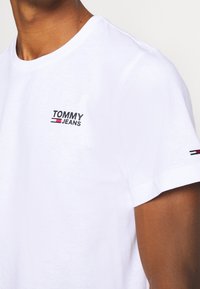 Vit bomull t-shirt med en svart och röd broderad "TOMMY JEANS" logotyp på bröstet och små trikoloraccenter på ärmen.