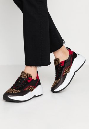 Legs in black cropped pants wearing chunky sneakers with leopard print, red and black panels, and thick white soles on a plain floor.