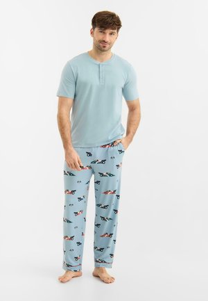 Mand stående barfodet iført lyseblå kortærmet henley og blå pyjamasbukser med racerbils- og ternet flagmønster.