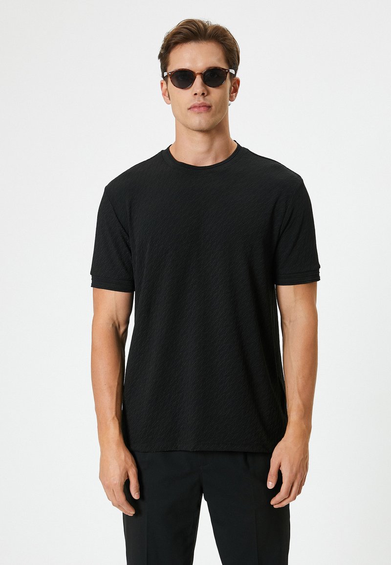 Sort t-shirt med kortærmer, der har et struktureret mønster, rund halsudskæring og løs pasform. Modellen bærer runde solbriller.