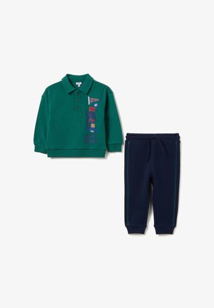 Grünes Baumwoll-Polo mit kurzen Ärmeln, das farbenfrohe Textmuster aufweist; kombiniert mit marineblauen Sweatpants mit grünen Seitenstreifen.