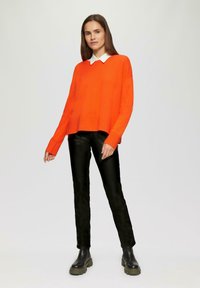Helles orange gestricktes Sweatshirt mit rundem Ausschnitt, langen Ärmeln und gerippten Bündchen, kombiniert mit schmalen schwarzen Hosen und klobigen schwarzen Stiefeln.