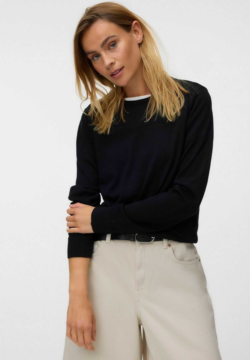 Femme aux cheveux blonds portant un haut noir à manches longues et un pantalon beige clair, debout avec un bras croisé sur son corps.