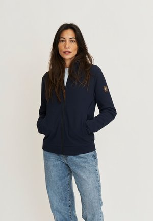 Vrouw met lang bruin haar, gekleed in een marineblauwe trui met ritssluiting en lichtblauwe spijkerbroek, staand met handen in de zakken tegen een effen achtergrond.