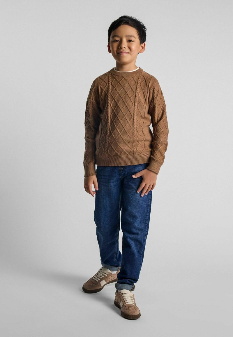Maglione lavorato a maglia marrone con motivi a diamante, jeans blu e sneakers marrone chiaro. Il maglione ha un collo rotondo e polsini a coste.