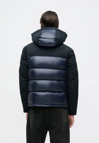 Doudoune noire et marine, dotée d'une capuche, avec un design matelassé horizontal au dos et une texture lisse et brillante.