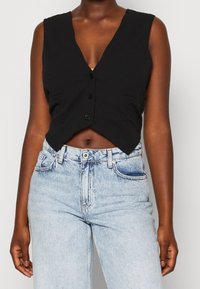 Mörkhyad person som bär en ärmlös svart knäppt crop top och ljusblå högmidjade jeans mot en helt vit bakgrund.