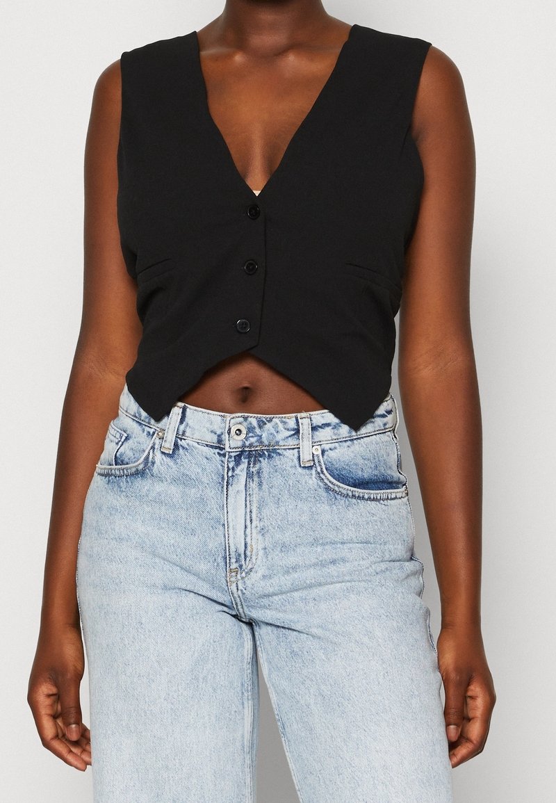 Mörkhyad person som bär en ärmlös svart knäppt crop top och ljusblå högmidjade jeans mot en helt vit bakgrund.