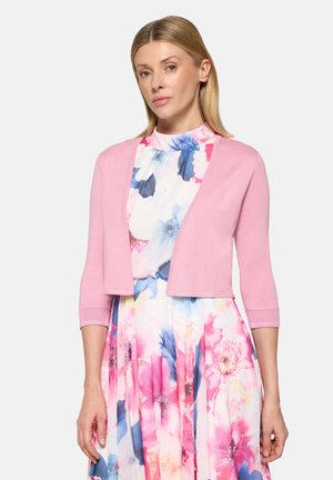 Woman wearing a pink cropped cardigan over a white dress with large pink and blue floral patterns, standing against a light background.