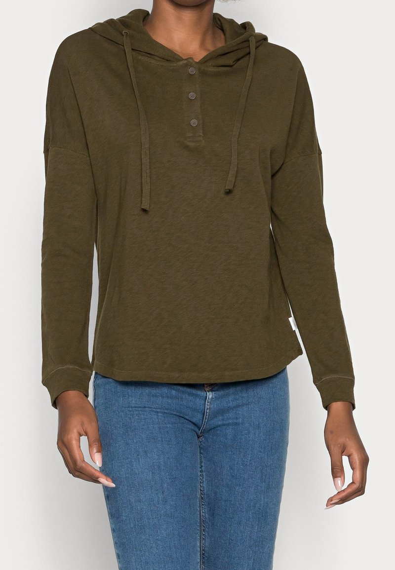 Personne portant un sweat à capuche kaki à manches longues avec des boutons et un jean bleu, debout devant un fond clair uni.