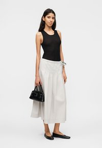 Γυναίκα που στέκεται, φοράει μαύρο αμάνικο τοπ, λευκή πλισέ midi φούστα, μαύρα μπαλαρίνια και κρατάει μια μικρή μαύρη τσάντα.