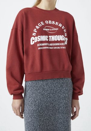 Osoba ubrana w czerwoną bluzę z białym napisem „Space Observer Cosmic Thoughts” i szarą teksturowaną spódnicę, stojąca na jednolitym tle.