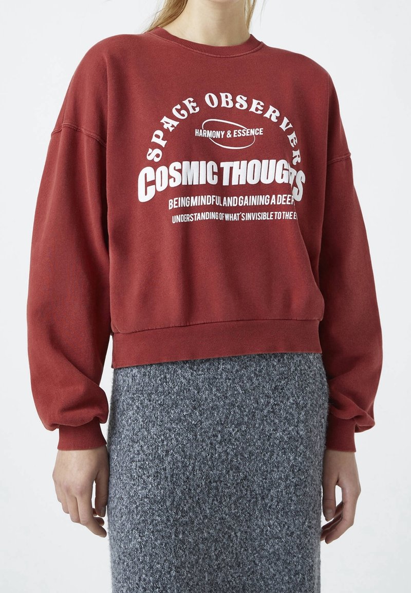 Personne portant un sweat-shirt rouge avec le texte blanc « Space Observer Cosmic Thoughts » et une jupe grise texturée, debout sur un fond uni.