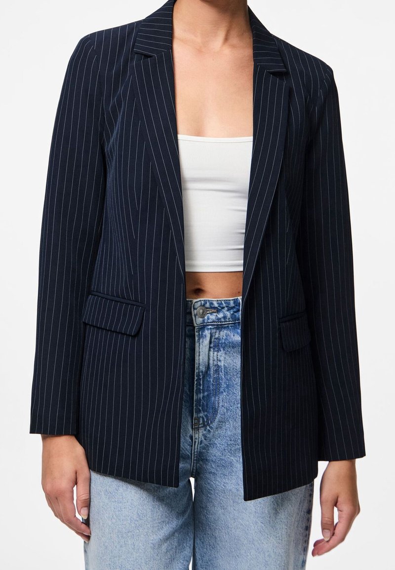 Femme portant un blazer bleu marine à fines rayures sur un top court blanc et un jean bleu clair taille haute, vue du milieu de la poitrine jusqu’aux hanches.