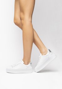 White sneakers with a smooth, glossy finish, round toe, raised platform sole, and gold-tone eyelets. Features a decorative loop at the heel.