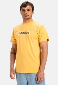 Yellow cotton t-shirt with short sleeves. Features a centered graphic logo in black and white with checkered detail. Classic cut and relaxed fit.