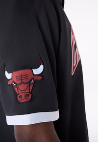 Μαύρη αθλητική φανέλα με στρογγυλή λαιμόκοψη, που διαθέτ ει κόκκινο και λευκό κεντημένο λογότυπο ταύρου στο αριστερό μανίκι και λευκές λεπτομέρειες στο τελείωμα.