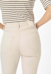 Beige high-waisted broeken met een gladde textuur, voorzien van twee achterzakken en een klein merklabel op de tailleband. Gestreepte top zichtbaar erboven.