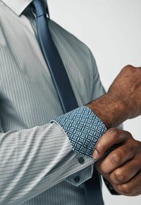 Langarmhemd met een lichtgrijze visgraatstructuur, contrasterende blauwe manchetten met een geometrisch ontwerp, gecombineerd met een gestructureerde blauwe stropdas.