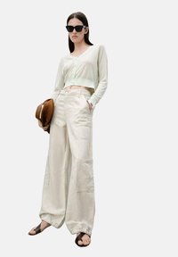 Ljusgrön kortärmad cardigan, breda beige cargobyxor med fickor och bruna sandaler. Modellen håller en brun hatt och bär svarta solglasögon.