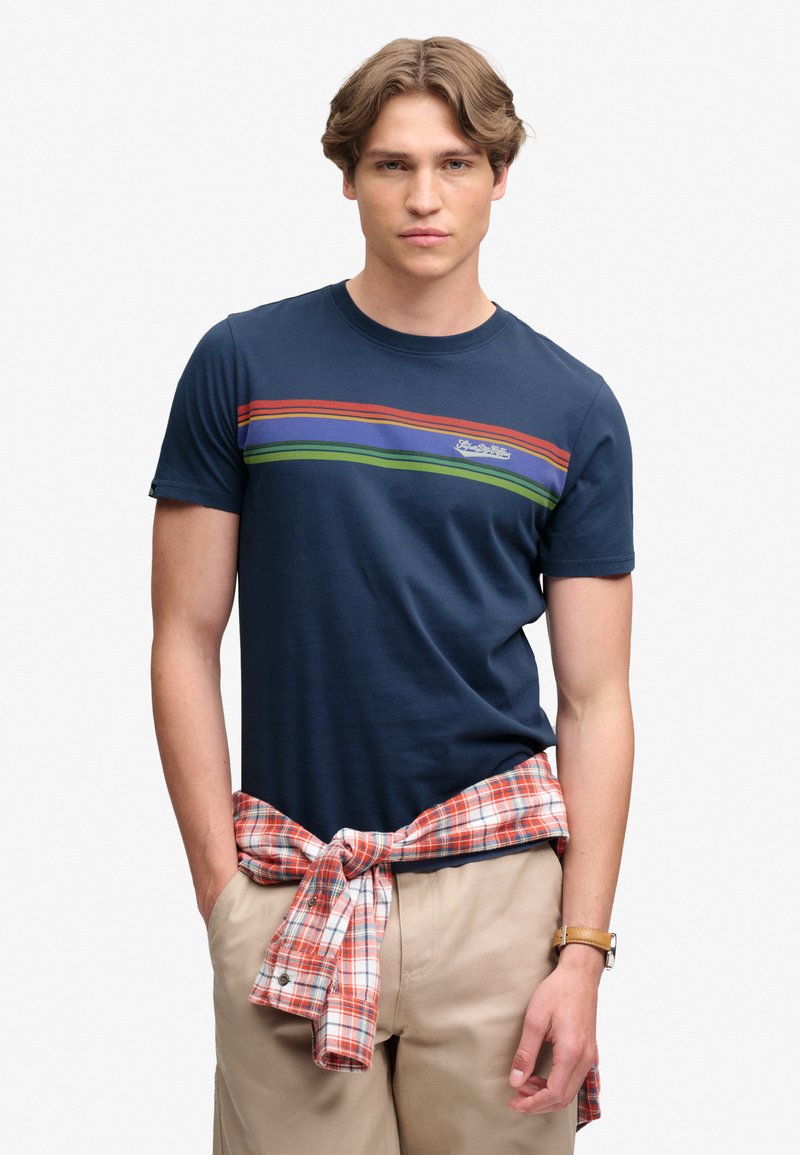 Joven vestido con una camiseta azul marino con rayas rojas, azules y verdes y una camisa de cuadros atada alrededor de su cintura con pantalones beige.