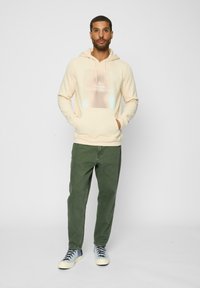 Cremefarvet hættebluse med et grafisk print på forsiden, parret med olivenfarvede bukser og sneakers med blå accenter. Teksturstof og afslappet pasform.
