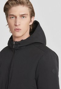 Giacca nera con cappuccio, dalla texture liscia, chiusura con zip e un piccolo logo sulla manica sinistra. Vestibilità sagomata e design minimal.