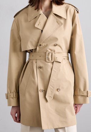 Kvinde iført en beige dobbeltradet trenchcoat med taljebælte og spænde, forlommer og manchetlommer, stående mod en ensfarvet baggrund.