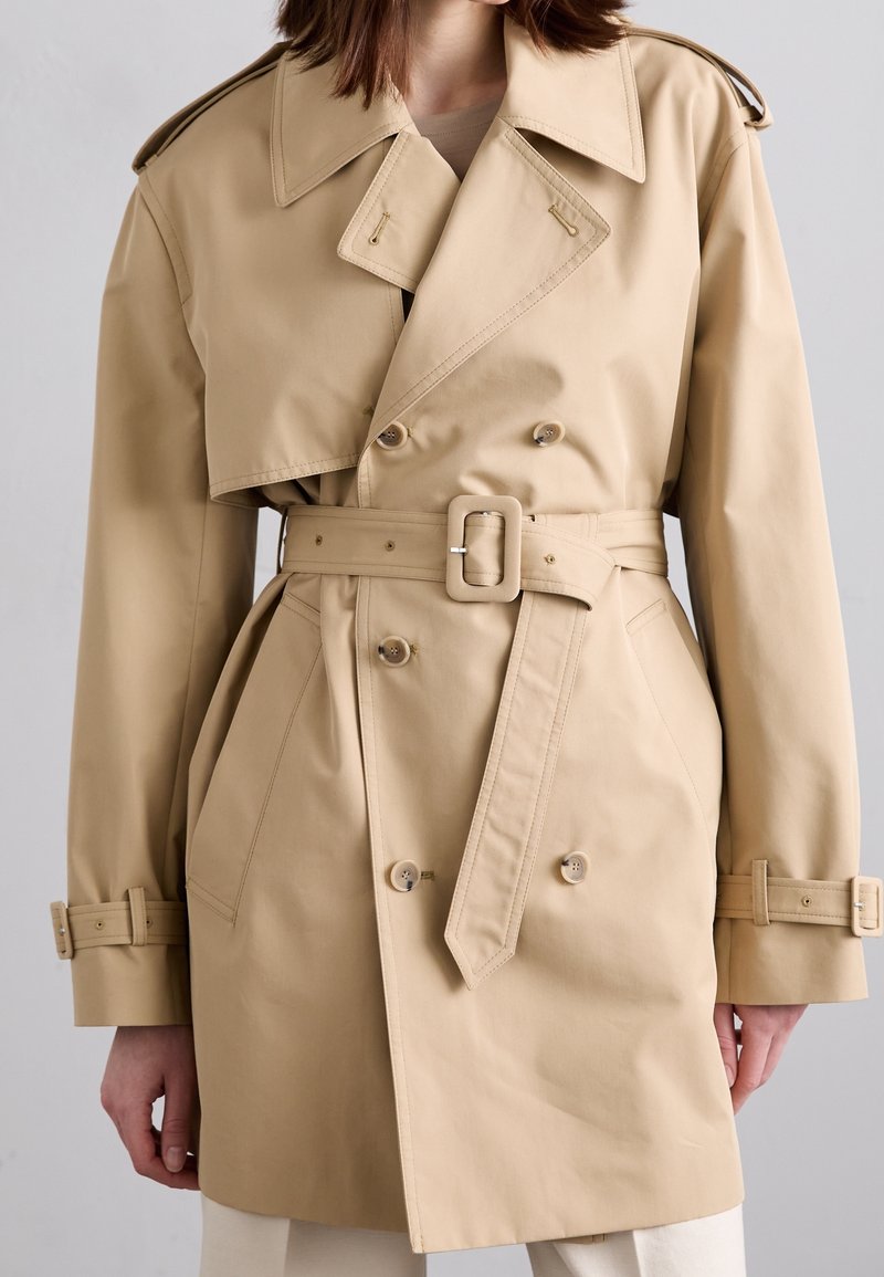 Femme portant un trench-coat beige croisé avec ceinture et boucle à la taille, poches avant et brides aux poignets, debout devant un fond uni.