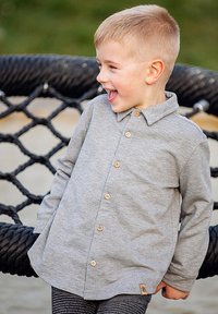 Szara koszula zapinana na guziki, wykonana z miękkiego materiału, z kołnierzykiem i drewnianymi guzikami. Projekt obejmuje długie rękawy i zaokrąglony dół.