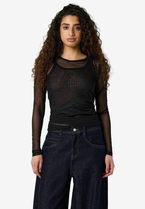 Femme aux longs cheveux bouclés portant un haut noir transparent à manches longues et un jean bleu foncé ample, debout devant un fond uni.