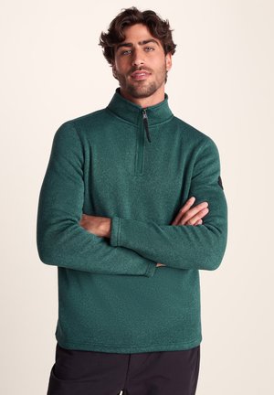 Green pullover made of textured fleece fabric, featuring a half-zip collar, long sleeves, and a logo patch on the left sleeve.