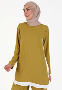 Long-sleeve tunic in olive green fabric, featuring a round neckline and side-slit white hem. Accompanied by a beige hijab.