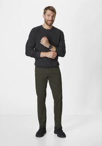 Dark gray knit sweater, olive green pants, black sneakers, and a watch with a dark dial. The individual stands confidently with arms crossed.